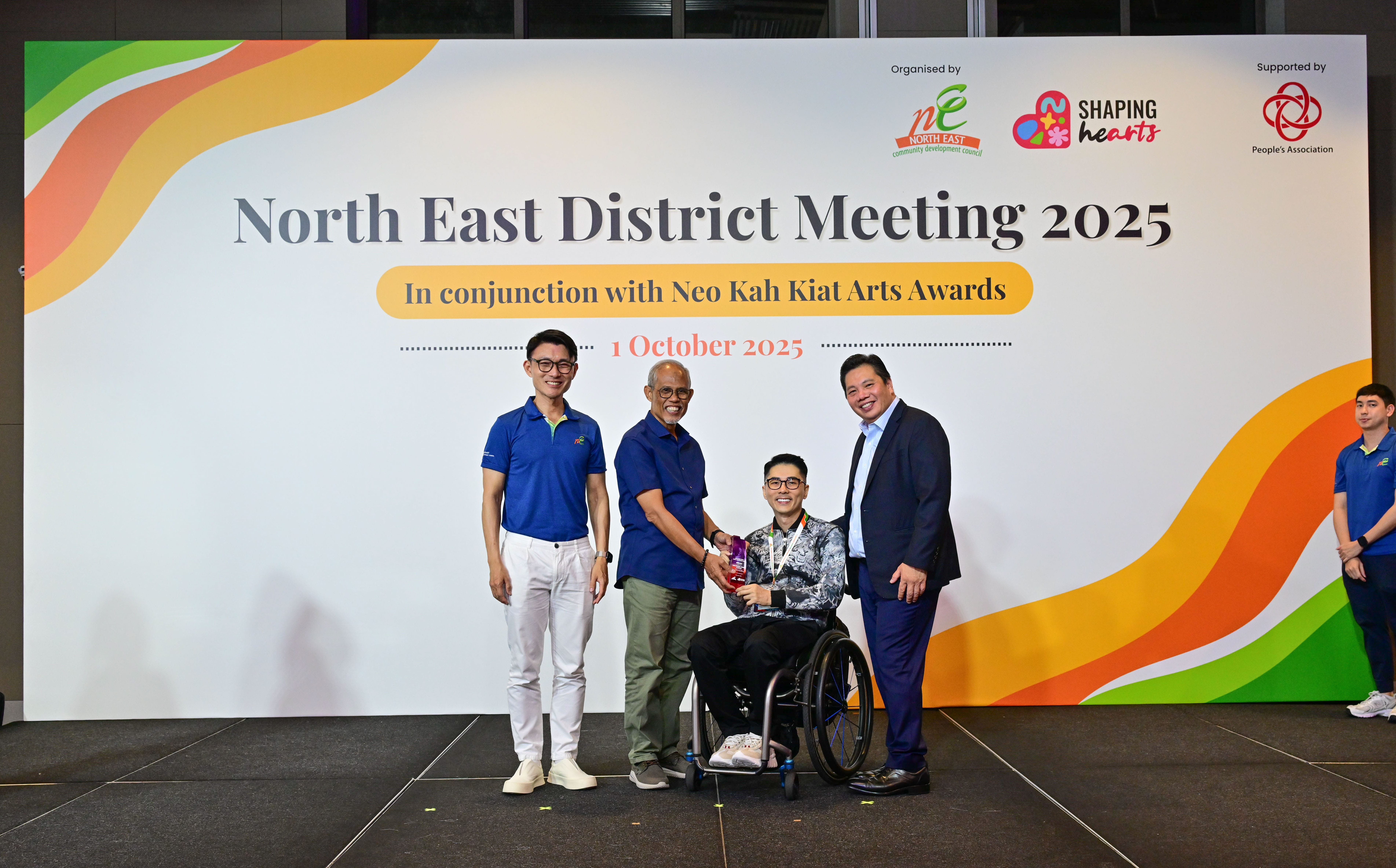 Award recipient standing on stage with the presenter, holding a acrylic plaque and posing for a group photo during the award ceremony, with a backdrop reading ‘North East District Meeting 2025 In conjunction with Neo Kah Kiat Arts Awards'